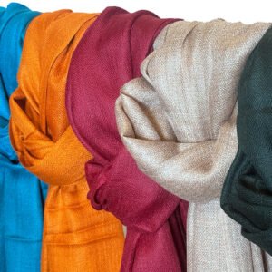 A row of richly textured shawls in various colors: black, teal, burnt orange, deep maroon, light beige, and dark green, draped over a bar.